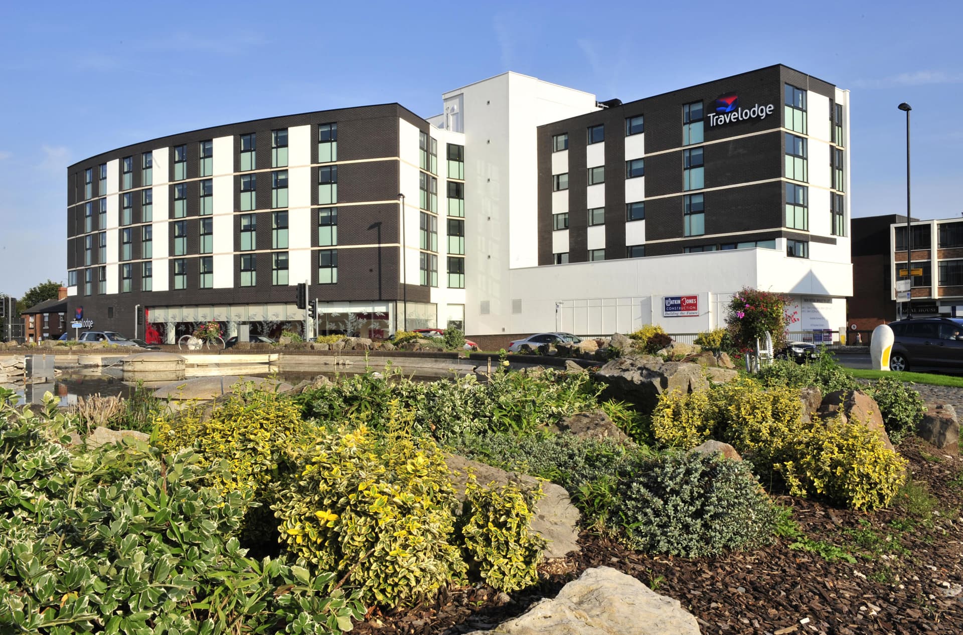 Large Travelodge hotel with rocks and green foliage in the foreground.